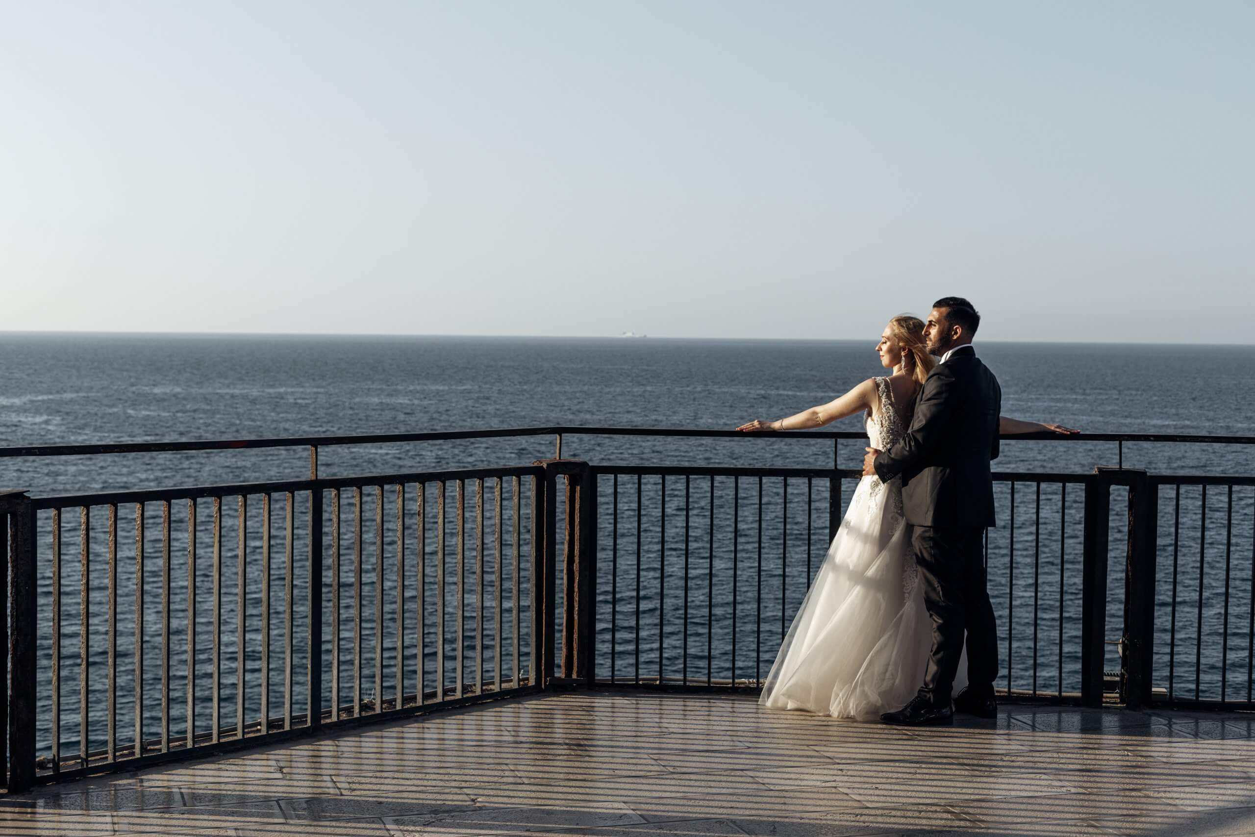 Side view of wedding couple standing at railing of embankment, enjoying view of Adriatic sea. Young woman bride with long wavy fair hair wearing long white wedding dress, hugging with groom in suit.